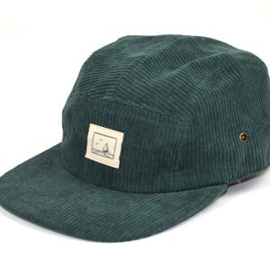 Op de afbeelding: Een donkergroene corduroy five-panel cap met een platte klep. De cap heeft een klein rechthoekig embleem met een zeilboot illustratie. Twee kleine metalen oogjes zitten aan de zijkant.