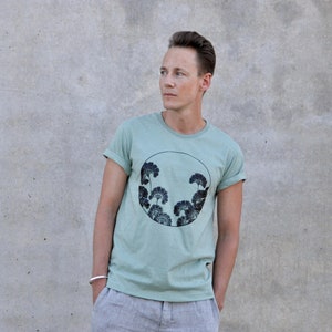 May include: A light green t-shirt with a black and white circular graphic of stylized trees. The graphic is centered on the shirt.
