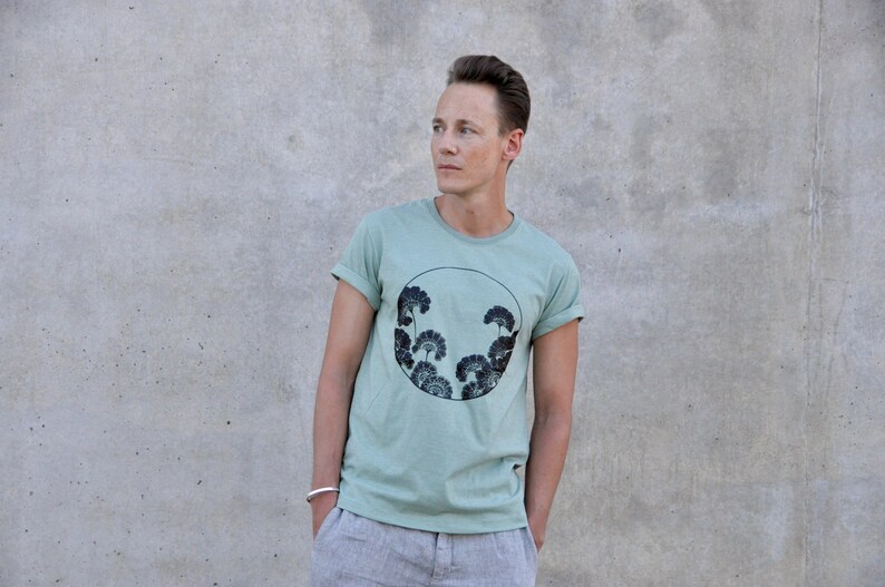 May include: A light green t-shirt with a black and white circular graphic of stylized trees. The graphic is centered on the shirt.