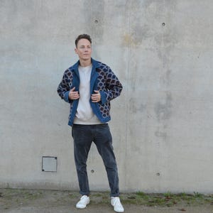 Peut inclure: Un homme portant une veste &agrave; motifs bleus avec une chemise blanche et un jean bleu fonc&eacute;. Il se tient devant un mur en b&eacute;ton.
