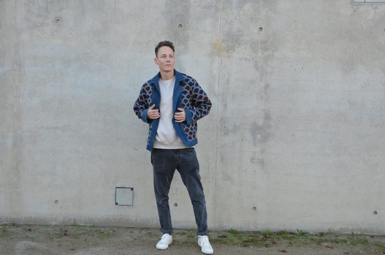 Peut inclure: Un homme portant une veste &agrave; motifs bleus avec une chemise blanche et un jean bleu fonc&eacute;. Il se tient devant un mur en b&eacute;ton.