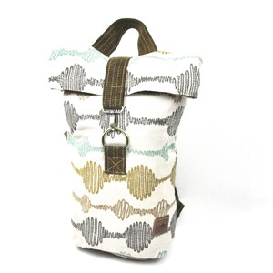 Peut inclure: Un sac à dos en toile blanche avec une sangle et une boucle en cuir marron. Le sac à dos présente un motif de lignes ondulées dans des tons de gris, vert et or.