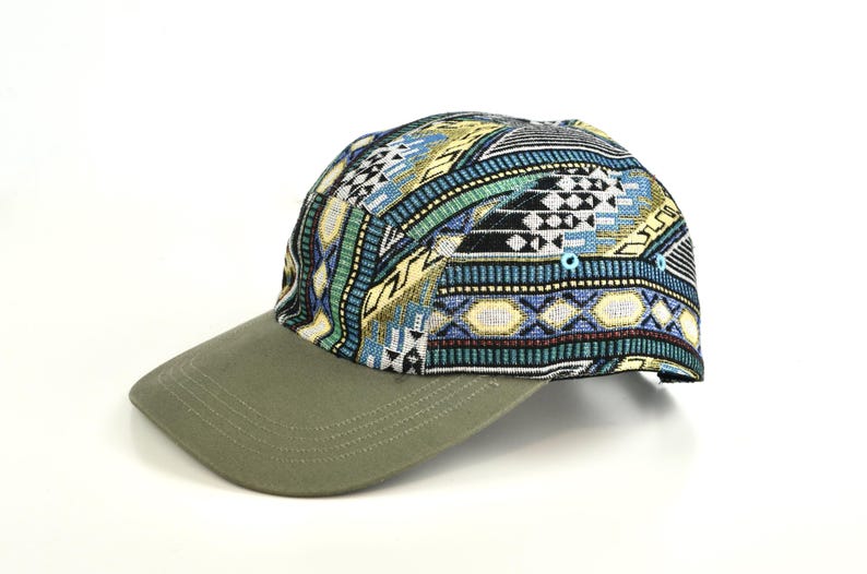 May include: A baseball cap with a patterned top and a solid green brim. The top of the cap features a colorful geometric design in shades of blue, yellow, black, and white. The brim is a solid olive green color.