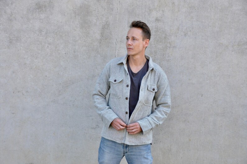 Puede incluir: Un hombre con una camisa de franela gris con una camiseta gris oscuro debajo y pantalones vaqueros azules. Est&aacute; de pie frente a una pared de hormig&oacute;n gris.