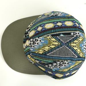 May include: A flat-brimmed cap with a patterned crown. The crown features a geometric design in blue, green, yellow, and black. The brim is a solid olive green color. This hat is a stylish accessory.