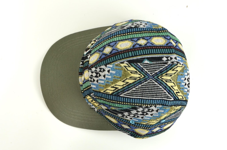 May include: A flat-brimmed cap with a patterned crown. The crown features a geometric design in blue, green, yellow, and black. The brim is a solid olive green color. This hat is a stylish accessory.