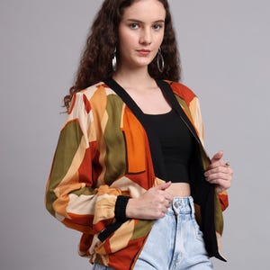 Könnte beinhalten: Eine Bomberjacke mit geometrischem Muster in Orange, Grün und Beige. Die Jacke hat schwarze Verzierungen und Bündchen und wird mit einem schwarzen Top und hellblauen Jeans kombiniert. Die Jacke ist offen, wodurch das schwarze Top sichtbar wird.