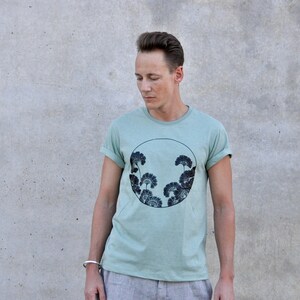 May include: A light green t-shirt with a black and white graphic of stylized trees in a circle. The graphic is centered on the shirt.