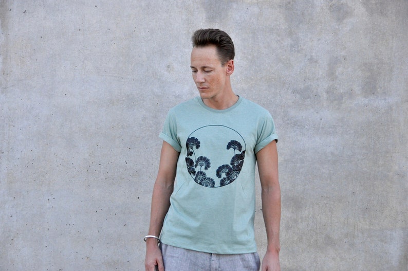 May include: A light green t-shirt with a black and white graphic of stylized trees in a circle. The graphic is centered on the shirt.