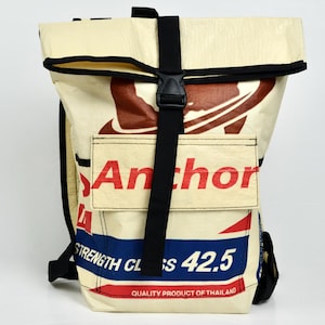 Könnte beinhalten: Ein Rolltop-Rucksack aus cremefarbenem Material mit schwarzen Riemen und einer Kunststoffschnalle. Die Vorderseite zeigt roten und blauen grafischen Text, darunter das Wort Anchor, den Schriftzug Strength Class 42.5 und Quality Product of Thailand.