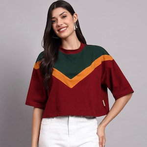 Puede incluir: Un top corto burdeos de manga corta con diseño de bloques de color. La parte superior presenta un patrón de chevron verde y amarillo en el pecho y los hombros. Tiene cuello redondo y una pequeña etiqueta en el dobladillo.