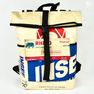 Op de afbeelding: Crèmekleurige rugzak met zwarte bandjes en een gespsluiting. De tas heeft blauwe, rode en witte bedrukte afbeeldingen, waaronder het woord "INSEE" en "RHINO PORTLAND". De tas heeft een handvat aan de bovenkant en zijvakken.