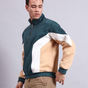 Könnte beinhalten: Ein farbblockierter Sweatshirt-Reißverschluss in Teal, Weiß und Beige. Das Sweatshirt hat einen hohen Kragen und einen Frontreißverschluss. Die Ärmel und der untere Saum sind teal. Das Model trägt eine beige Hose.