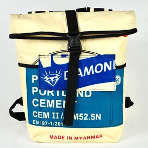 Op de afbeelding: Crèmekleurige rugzak met zwarte accenten en een zwarte gespsluiting. De voorkant heeft een blauw en wit label met "DIAMOND" en "PORTLAND CEMENT". De onderkant van de tas leest "MADE IN MYANMAR".