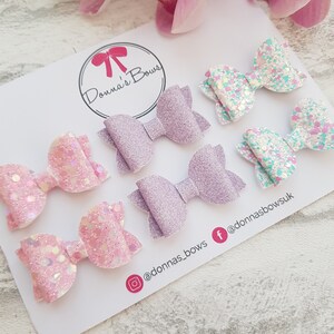 Peut inclure: Ensemble de six n&oelig;uds en paillettes pour cheveux en rose, lavande et turquoise. Les n&oelig;uds sont dispos&eacute;s en deux rang&eacute;es de trois. Les n&oelig;uds sont sur un fond blanc avec le texte "Donna's Bows" et les poign&eacute;es des m&eacute;dias sociaux.