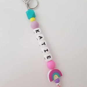 May include: A keyring featuring a silver heart-shaped clasp and ring, followed by a sequence of colourful beads including a teal geometric bead, a yellow bead, a lavender bead, white letter beads spelling KATIE, a pink bead, and a rainbow charm.