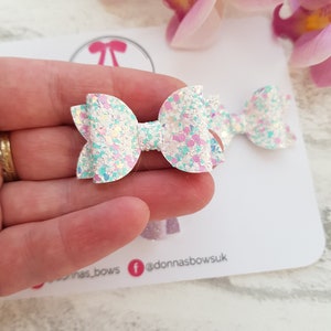 Peut inclure: Deux n&oelig;uds en cheveux blancs &agrave; paillettes avec des accents roses et bleus. Les n&oelig;uds sont tenus dans une main.
