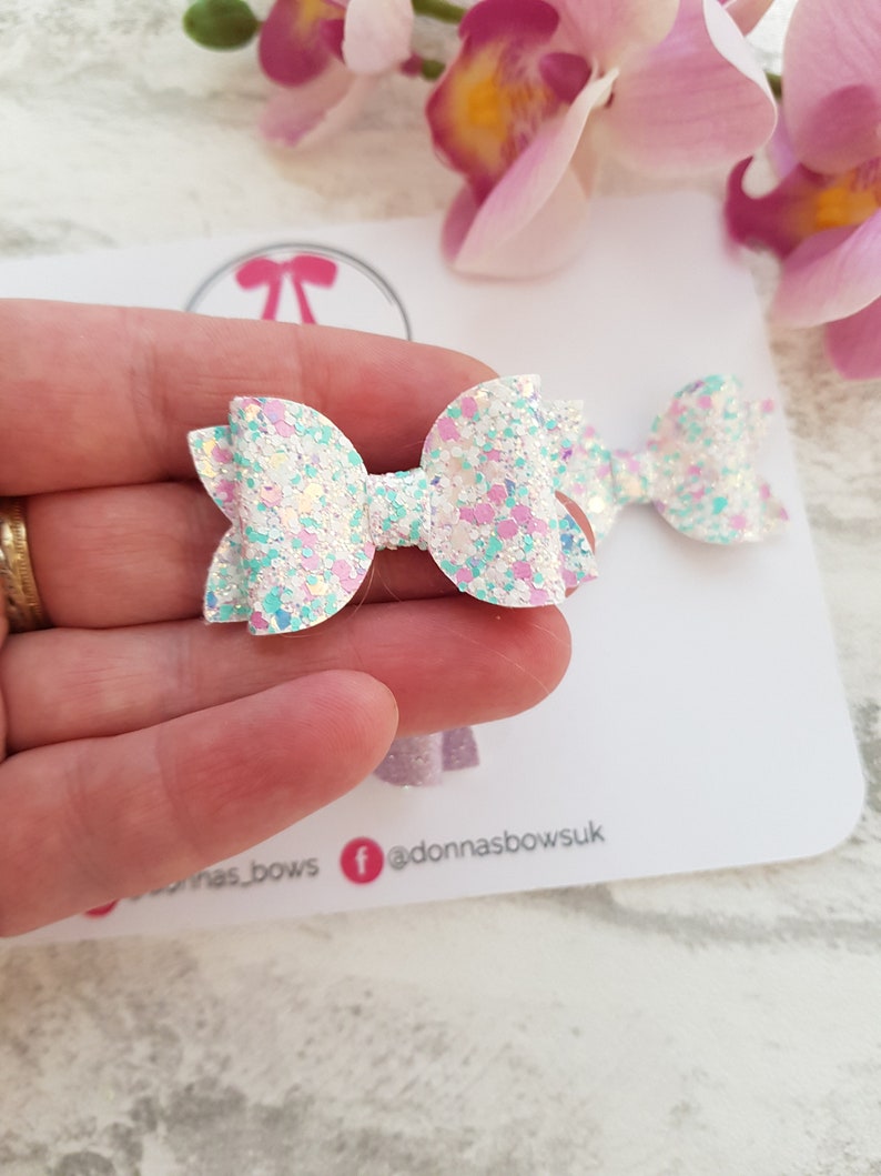 Peut inclure: Deux n&oelig;uds en cheveux blancs &agrave; paillettes avec des accents roses et bleus. Les n&oelig;uds sont tenus dans une main.