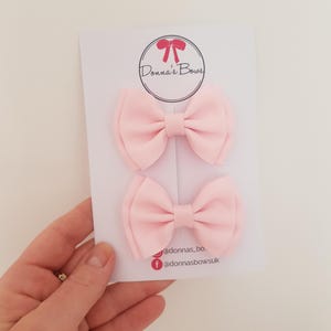 May include: Two light pink hair bows on a white card. The bows are made of a ribbed fabric and are tied in a classic bow shape. The card has the text "Donna's Bows" in a circle with a bow graphic.