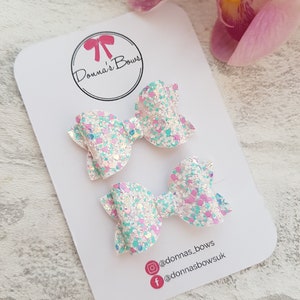 Peut inclure: Deux petits n&oelig;uds pour cheveux en paillettes blanches avec des accents de paillettes roses et bleues. Les n&oelig;uds sont sur une carte blanche avec le texte "Donna's Bows" et des poign&eacute;es de m&eacute;dias sociaux.