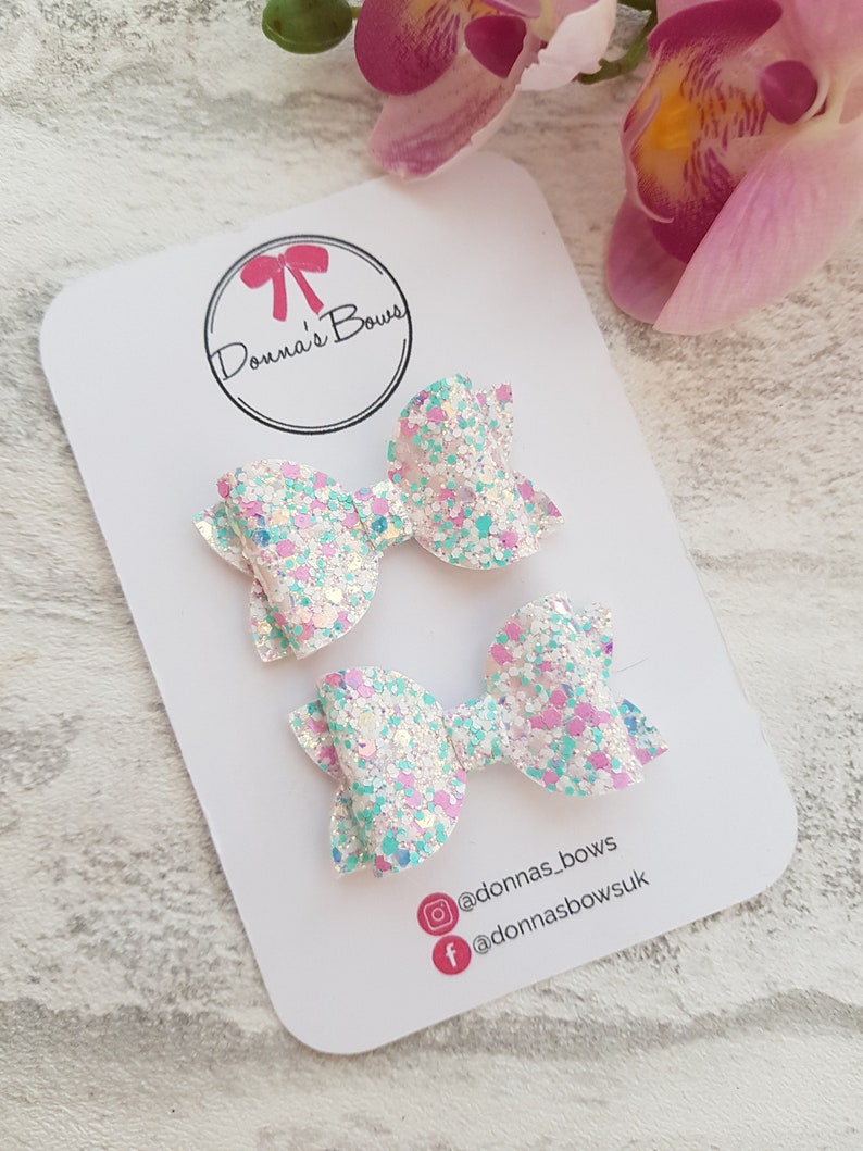 Peut inclure: Deux petits n&oelig;uds pour cheveux en paillettes blanches avec des accents de paillettes roses et bleues. Les n&oelig;uds sont sur une carte blanche avec le texte "Donna's Bows" et des poign&eacute;es de m&eacute;dias sociaux.