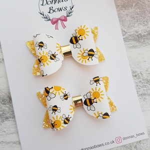 May include: Two white hair bows featuring a bee and sun design, gold glitter details, and gold metal clips. The bows are displayed on a card with the text "Donna's Bows" and social media information.
