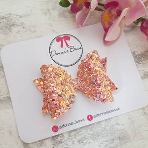 May include: A pair of pink and gold glitter hair bows with a scalloped edge. The bows are on a white card with the text "Donna's Bows" and social media handles.