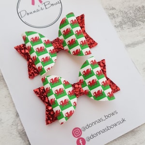 May include: Two red and green glitter hair bows with a Welsh dragon pattern. The bows are on a white card with the text "Donna's Bows" and social media handles @donnas_bows and @donnasbowsuk.
