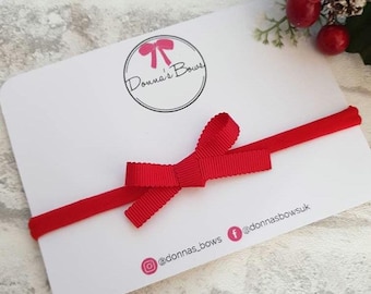 Puede incluir: Una diadema de cinta roja con un pequeño lazo rojo. La diadema está sobre una tarjeta blanca con el texto "Donna's Bows" y las manijas de las redes sociales.