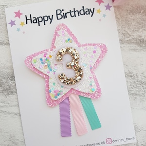 May include: A pink and white star-shaped hair clip with the number 3 in gold glitter. The clip is on a white card with the words "Happy Birthday" and colorful star accents. Three pastel ribbons hang below the star.