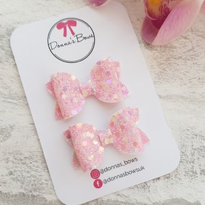 Peut inclure: Deux n&oelig;uds pour cheveux en paillettes roses avec des accents iris&eacute;s. Les n&oelig;uds sont pr&eacute;sent&eacute;s sur une carte blanche avec le texte "Donna's Bows" et des poign&eacute;es de m&eacute;dias sociaux.