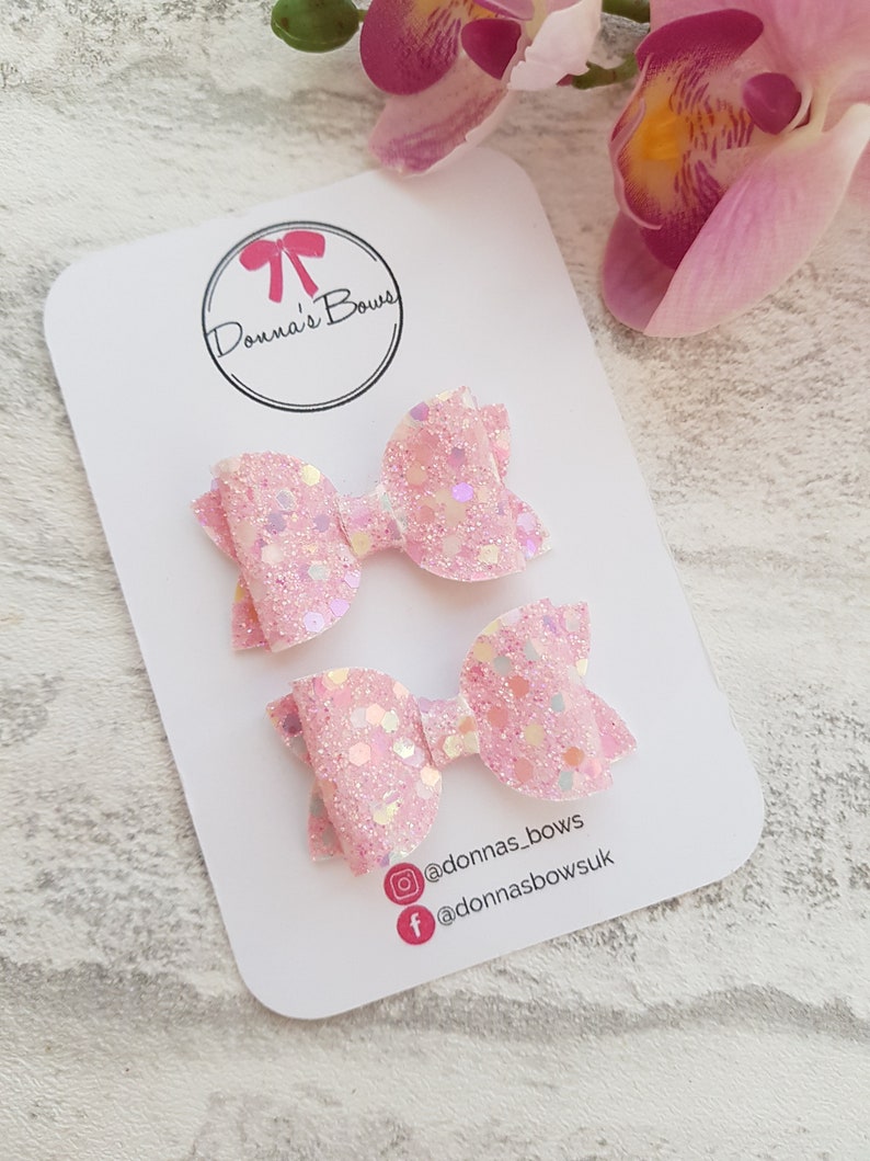Peut inclure: Deux n&oelig;uds pour cheveux en paillettes roses avec des accents iris&eacute;s. Les n&oelig;uds sont pr&eacute;sent&eacute;s sur une carte blanche avec le texte "Donna's Bows" et des poign&eacute;es de m&eacute;dias sociaux.