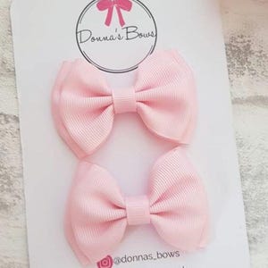May include: Two light pink hair bows on a white card with the text "Donna's Bows" in a circle. The bows are made of a ribbed fabric. The card also includes social media handles.