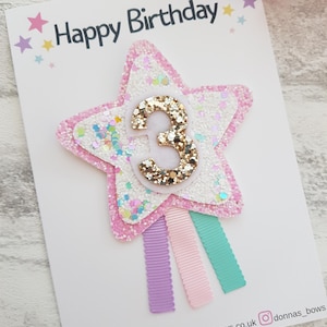 May include: A birthday card with the words "Happy Birthday" at the top. The card features a star-shaped embellishment with the number "3" in gold glitter. The star is surrounded by pink and white glitter. Three colorful ribbons are attached.