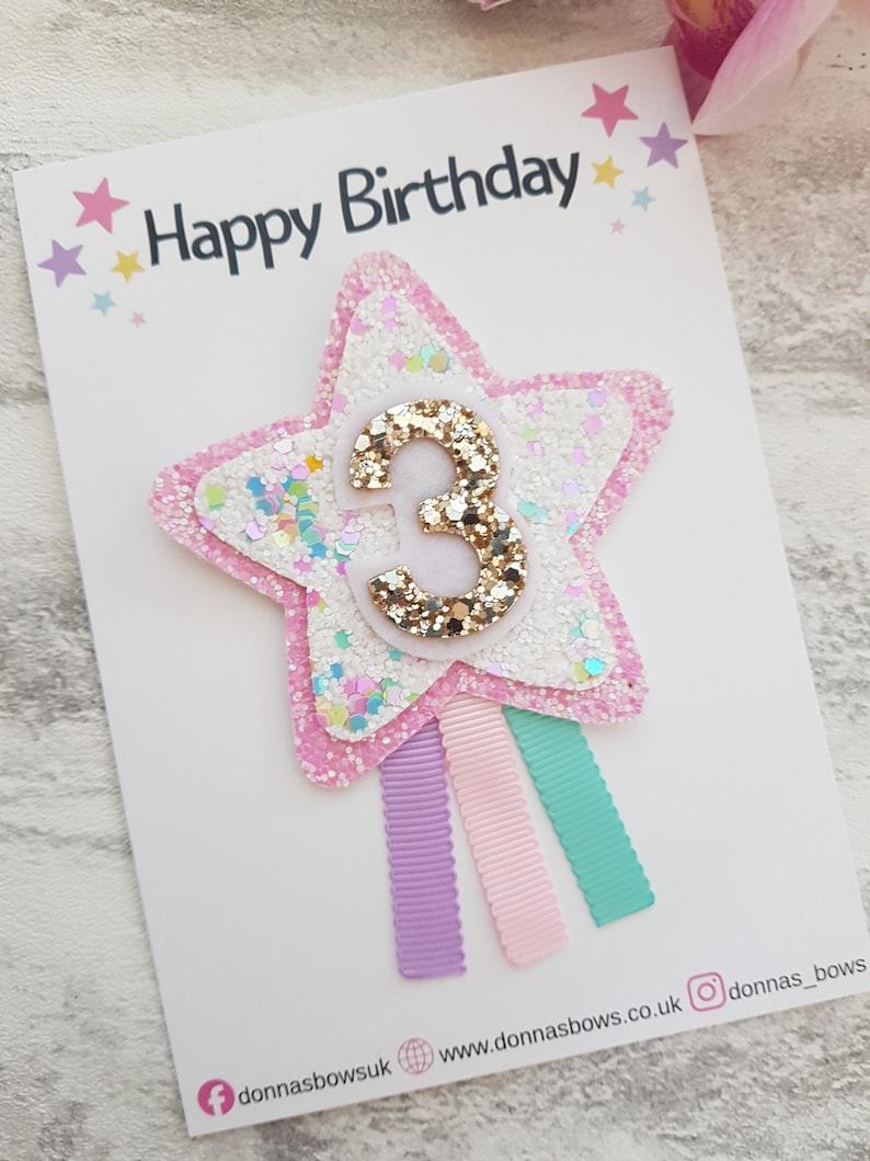 May include: A birthday card with the words "Happy Birthday" at the top. The card features a star-shaped embellishment with the number "3" in gold glitter. The star is surrounded by pink and white glitter. Three colorful ribbons are attached.