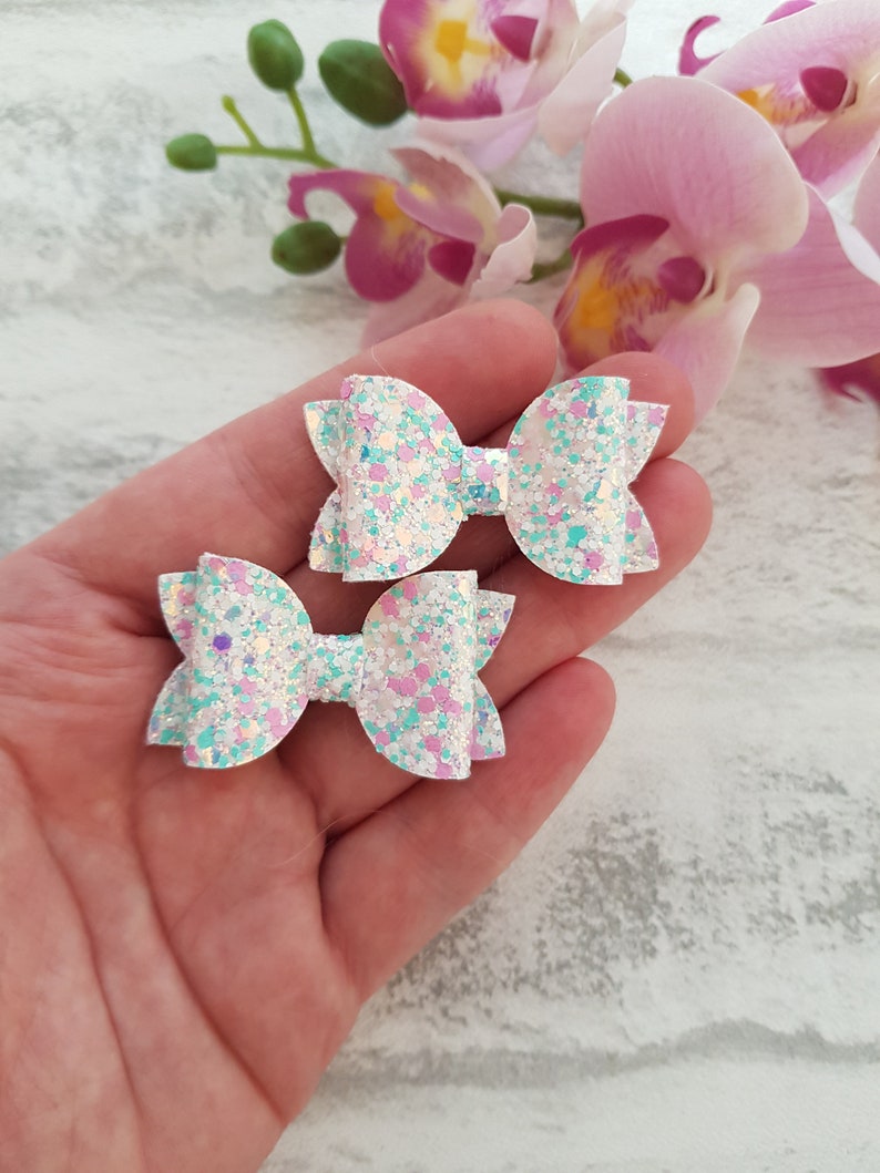 Peut inclure: Deux n&oelig;uds pour cheveux en paillettes blanches avec des paillettes iris&eacute;es et bleu pastel et rose. Les n&oelig;uds sont nou&eacute;s en un simple n&oelig;ud.
