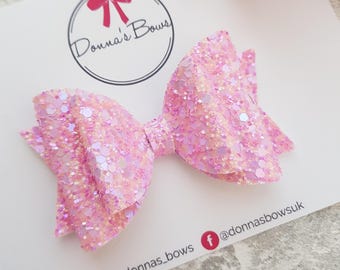 May include: A pink glitter hair bow with a white center. The bow is on a white card with the text "Donna's Bows" and social media handles.