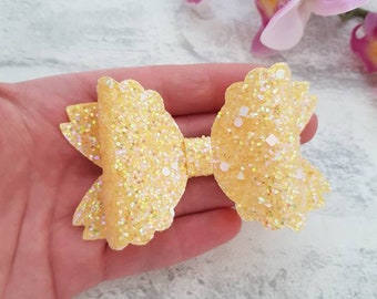 May include: A yellow glitter bow with a scalloped edge. The bow is made of a soft, sparkly material and is perfect for adding a touch of whimsy to any outfit.