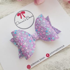 May include: A purple glitter hair bow with iridescent accents. The bow is on a white card with the text "Donna's Bows" and social media handles @donnas_bows and @donnasbowsuk.