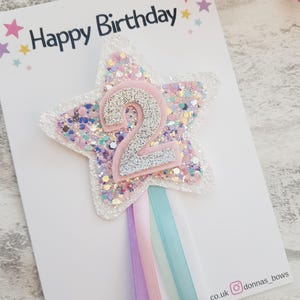 May include: A star-shaped birthday cake topper with the number 2 in silver glitter. The star is white with pink, purple, and silver glitter. The words "Happy Birthday" are printed at the top. Pastel ribbons hang from the bottom.