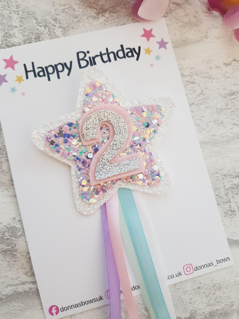 May include: A star-shaped birthday cake topper with the number 2 in silver glitter. The star is white with pink, purple, and silver glitter. The words "Happy Birthday" are printed at the top. Pastel ribbons hang from the bottom.