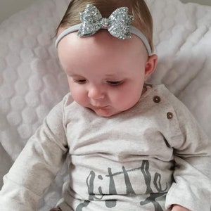 Peut inclure: Un b&eacute;b&eacute; portant un t-shirt gris &agrave; manches longues avec le texte "little" imprim&eacute; dessus. Le b&eacute;b&eacute; porte un bandeau argent&eacute; &agrave; paillettes avec un n&oelig;ud.