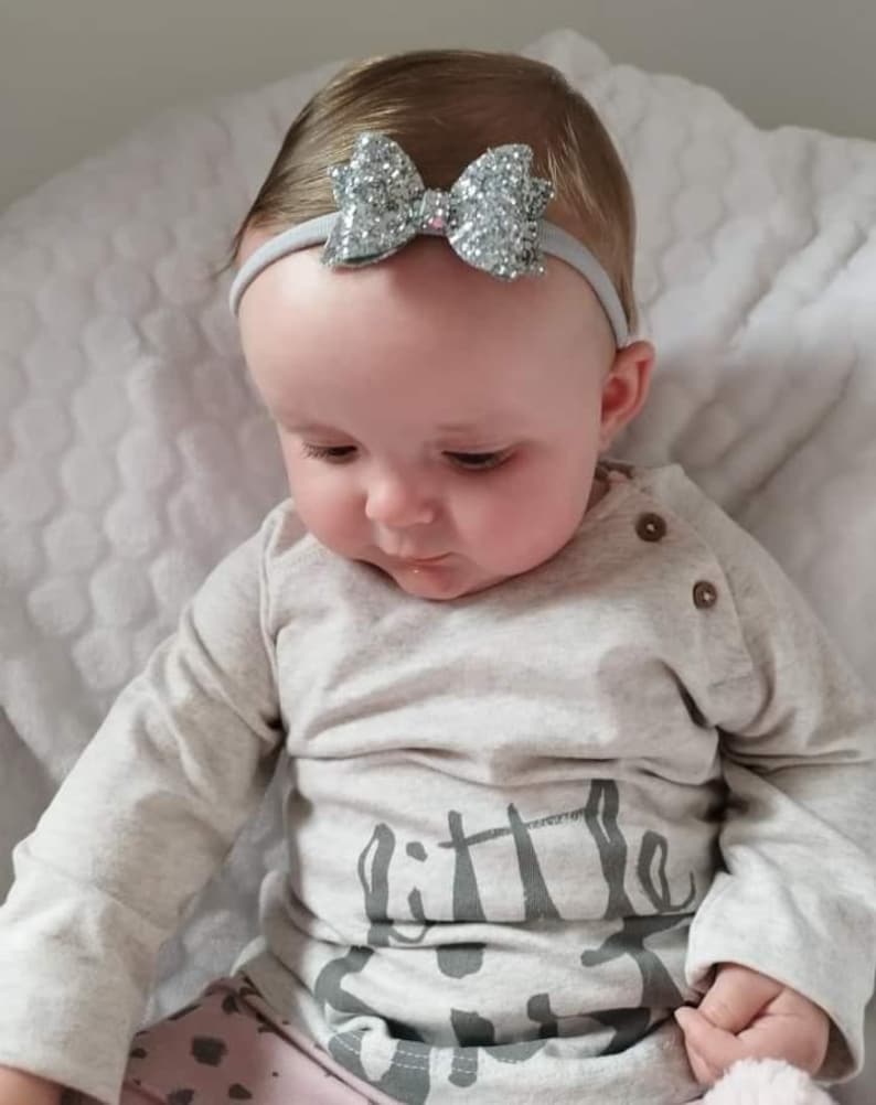 Peut inclure: Un b&eacute;b&eacute; portant un t-shirt gris &agrave; manches longues avec le texte "little" imprim&eacute; dessus. Le b&eacute;b&eacute; porte un bandeau argent&eacute; &agrave; paillettes avec un n&oelig;ud.