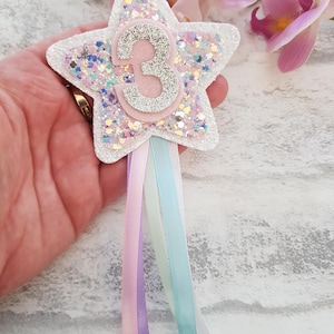 A star-shaped birthday badge featuring the chunky glitters. The rosette has pastel ribbons in pink, lilac, and turquoise. The star is layered with glitter and has a pink and silver numbers.