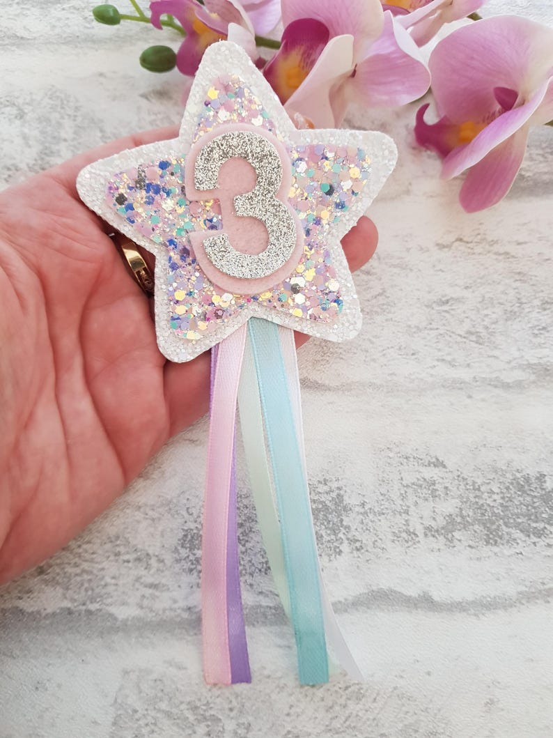 A star-shaped birthday badge featuring the chunky glitters. The rosette has pastel ribbons in pink, lilac, and turquoise. The star is layered with glitter and has a pink and silver numbers.