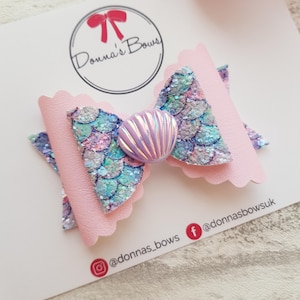 May include: A pink and blue mermaid-themed hair bow with a sparkly iridescent seashell in the centre. The bow is on a white card with the text "Donna's Bows" and social media handles.
