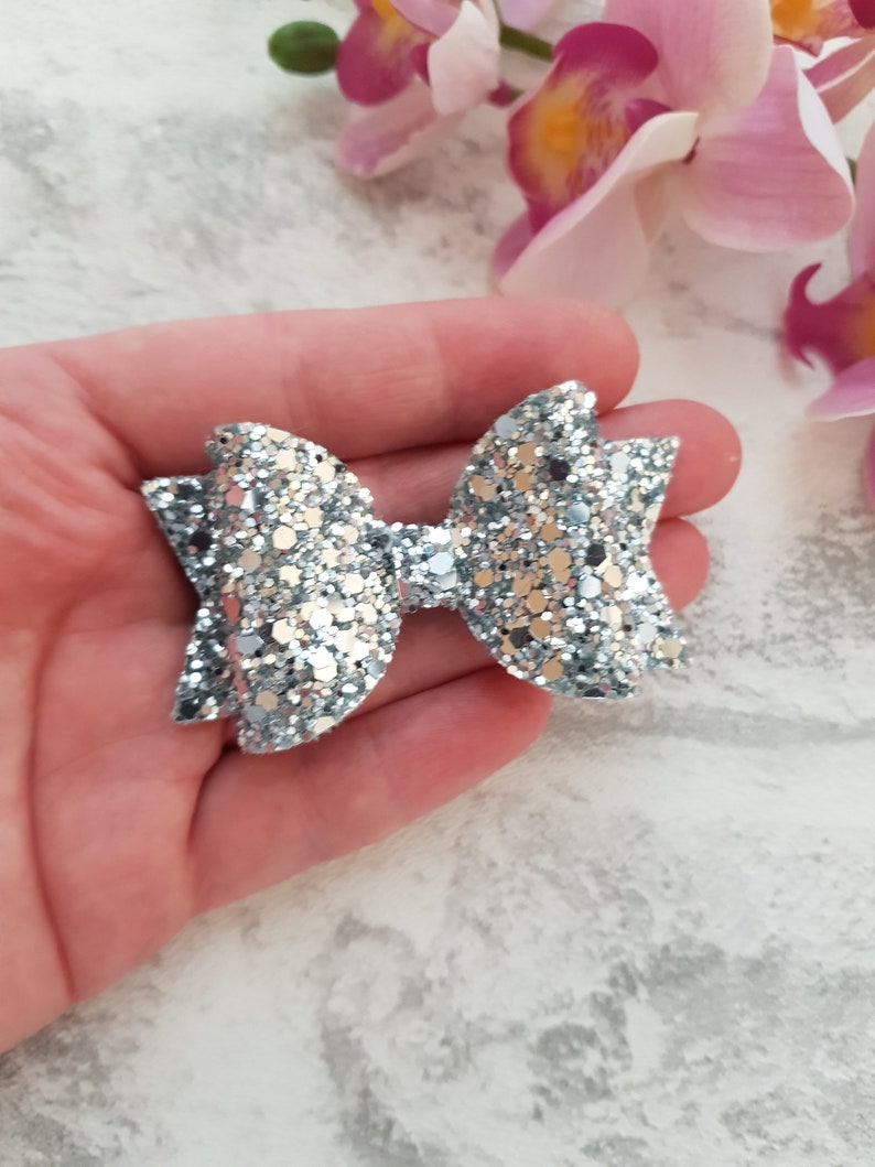 Peut inclure: Un n&oelig;ud en paillettes argent&eacute;es avec une boucle centrale. Le n&oelig;ud est tenu dans une main.