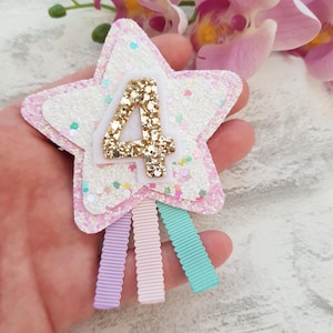 May include: A star-shaped hair clip with a glittery white and pink design. The number "4" is in gold glitter. Three pastel ribbons in lavender, pink, and teal hang from the bottom. The clip is likely for a child's birthday.
