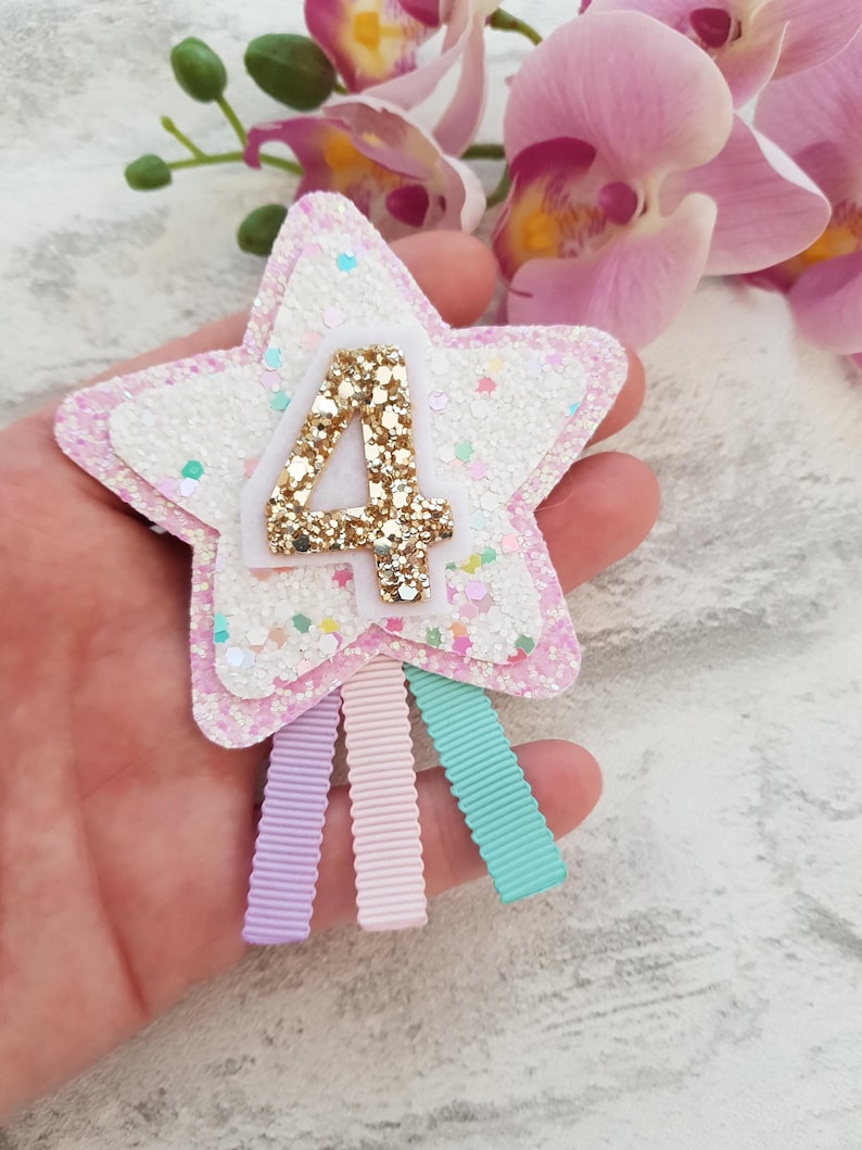 May include: A star-shaped hair clip with a glittery white and pink design. The number "4" is in gold glitter. Three pastel ribbons in lavender, pink, and teal hang from the bottom. The clip is likely for a child's birthday.