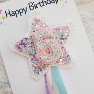 May include: A birthday card with the words "Happy Birthday" at the top. The card features a star-shaped decoration with the number 5 in silver glitter, surrounded by pink and iridescent glitter. Ribbons in pastel colors hang from the bottom.